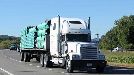 A white Peterbilt flatbed truck transporting industrial pipes on a highway