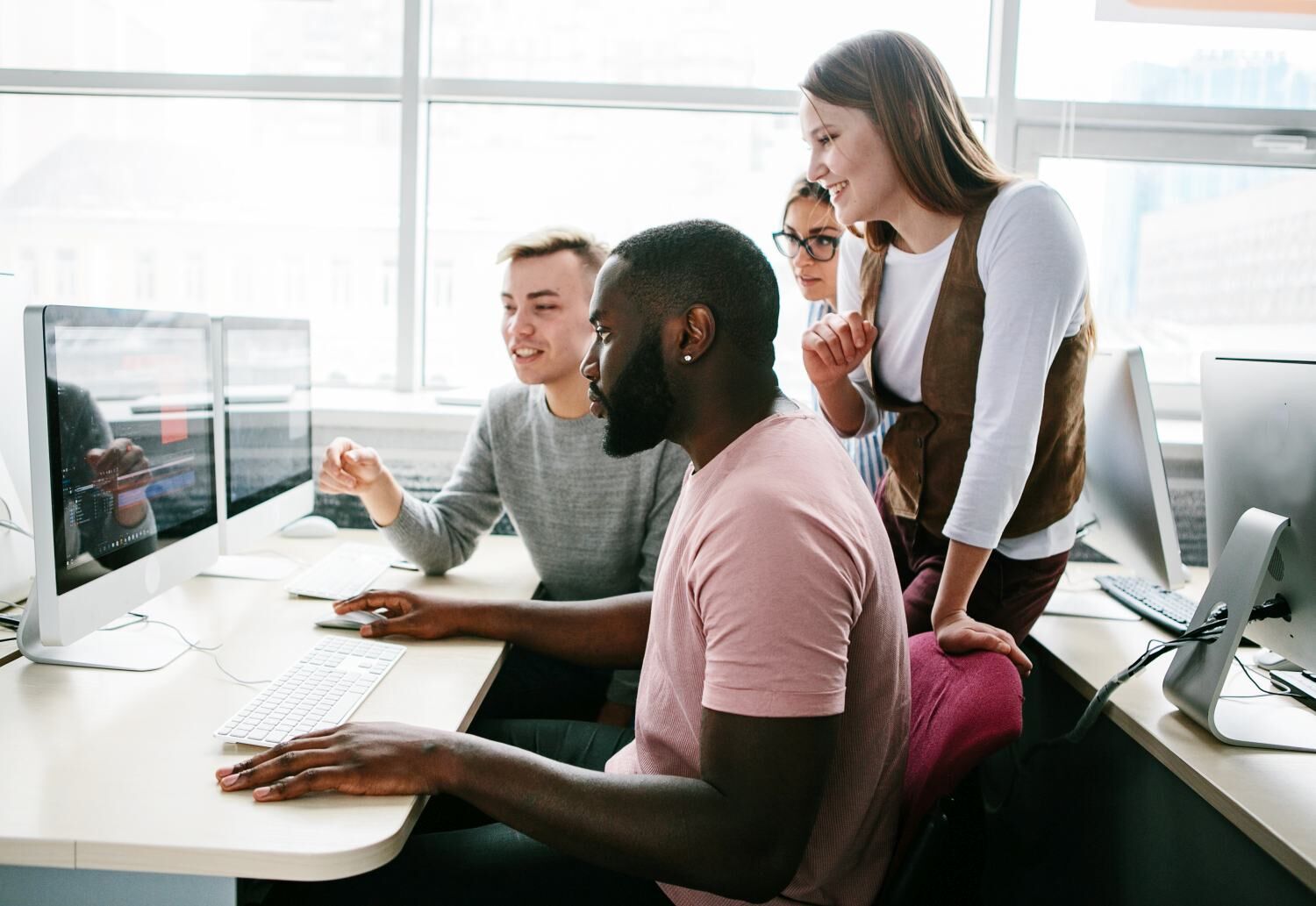 Group of young professionals collaborating and discussing work at computer stations in a bright office.